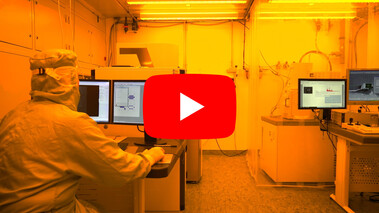 Screenshot eines Videos aus einem Reinraumlabor mit gelber Beleuchtung, Forscher an Computerarbeitsplätzen.